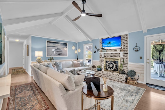 living room with lofted ceiling with beams, wood finished floors, wainscoting, a brick fireplace, and ceiling fan