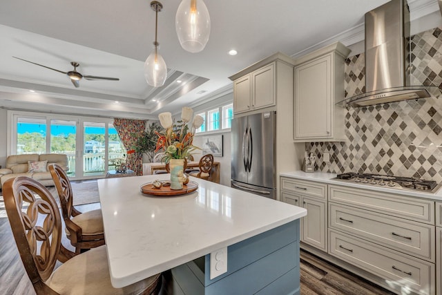 kitchen with light countertops, ornamental molding, a raised ceiling, wall chimney range hood, and appliances with stainless steel finishes