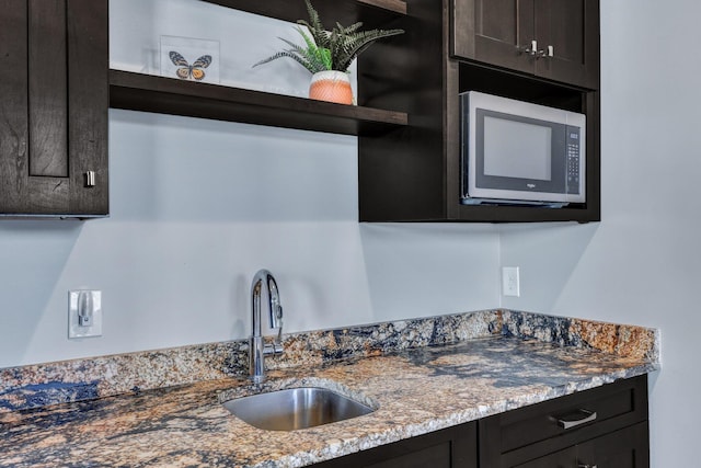 kitchen featuring open shelves, stainless steel microwave, a sink, light stone counters, and dark brown cabinets