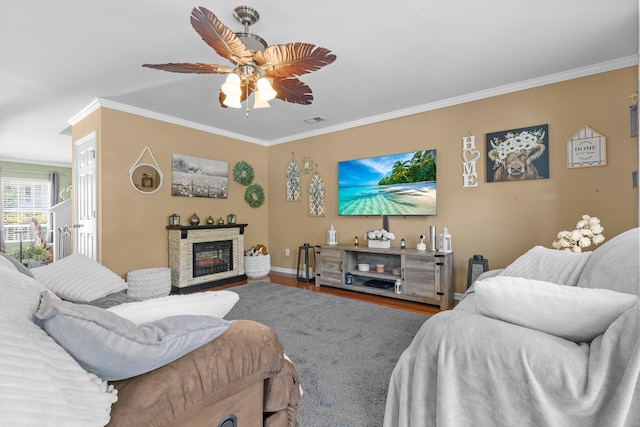 living area with ornamental molding, a stone fireplace, a ceiling fan, baseboards, and wood finished floors