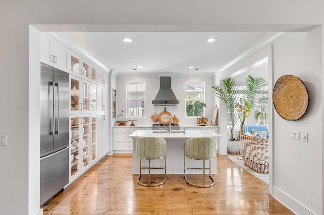 kitchen with light hardwood / wood-style floors, white cabinets, a center island with sink, wall chimney range hood, and stainless steel built in fridge