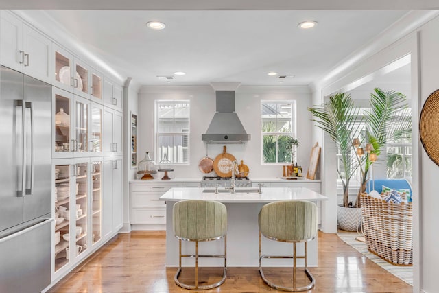 kitchen featuring white cabinets, built in refrigerator, a kitchen island with sink, light hardwood / wood-style flooring, and wall chimney range hood