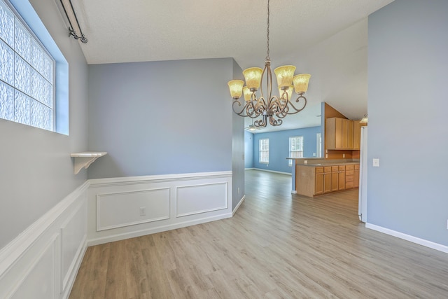 unfurnished dining area with a notable chandelier, light wood-style flooring, a textured ceiling, wainscoting, and vaulted ceiling