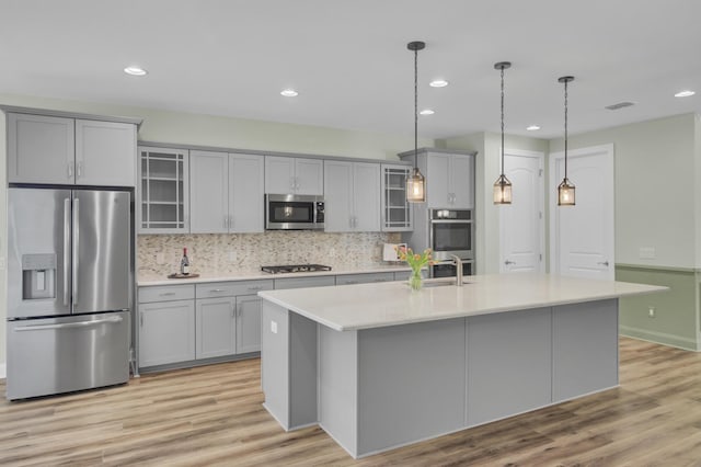 kitchen with decorative backsplash, appliances with stainless steel finishes, a kitchen island with sink, decorative light fixtures, and gray cabinets