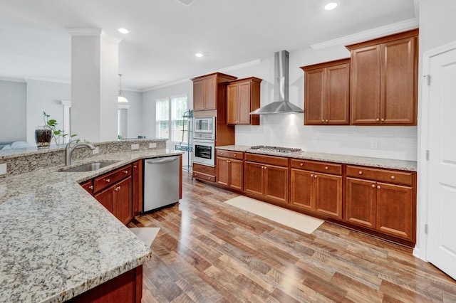 kitchen featuring light wood finished floors, wall chimney exhaust hood, appliances with stainless steel finishes, crown molding, and a sink