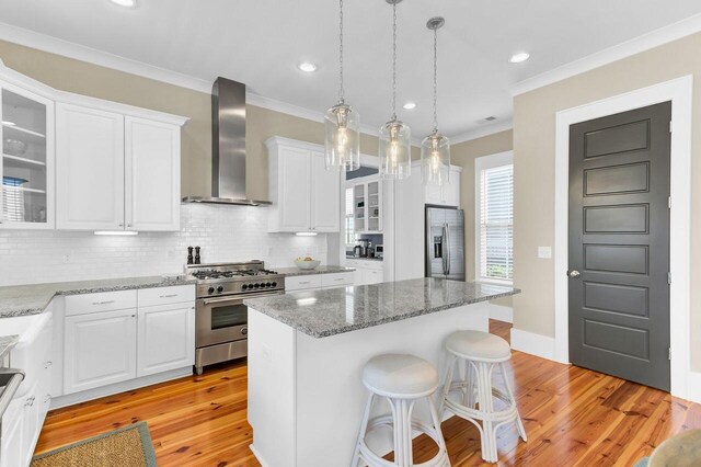 kitchen with ornamental molding, stainless steel appliances, a kitchen island, white cabinetry, and wall chimney range hood