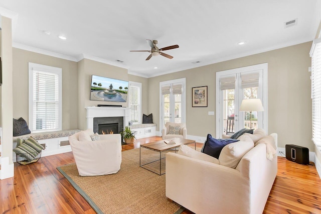 living area featuring crown molding, hardwood / wood-style flooring, a ceiling fan, a warm lit fireplace, and visible vents