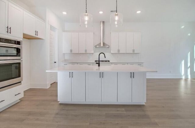 kitchen featuring a kitchen island with sink, pendant lighting, white cabinets, and wall chimney exhaust hood