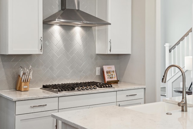 kitchen featuring light stone countertops, stainless steel gas cooktop, sink, wall chimney range hood, and white cabinetry