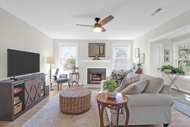 living room featuring visible vents, a fireplace with flush hearth, light wood-style floors, baseboards, and ceiling fan