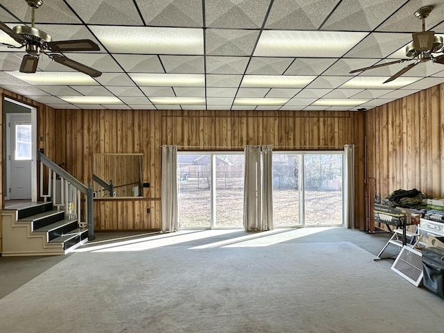 carpeted living room featuring ceiling fan, a paneled ceiling, and wooden walls