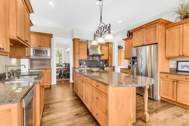 kitchen featuring pendant lighting, wall chimney range hood, a kitchen island with sink, stainless steel appliances, and dark stone counters