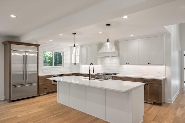 kitchen featuring premium range hood, a center island with sink, decorative light fixtures, light hardwood / wood-style floors, and stainless steel appliances