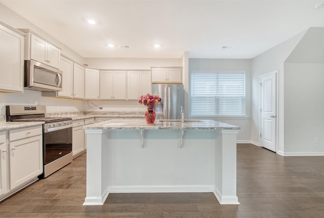 kitchen with light stone countertops, stainless steel appliances, a kitchen island with sink, white cabinets, and dark hardwood / wood-style floors