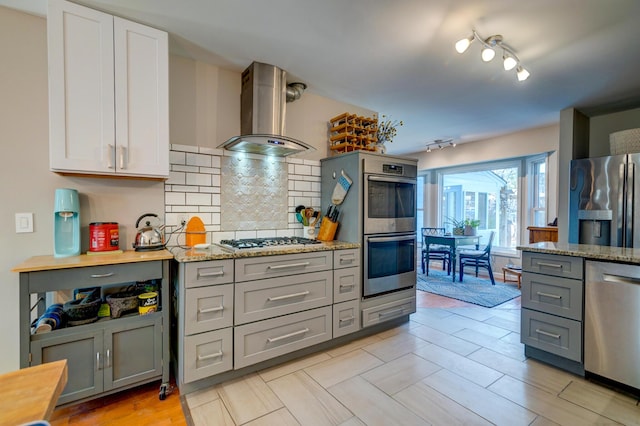 kitchen with backsplash, light stone countertops, gray cabinets, appliances with stainless steel finishes, and wall chimney exhaust hood