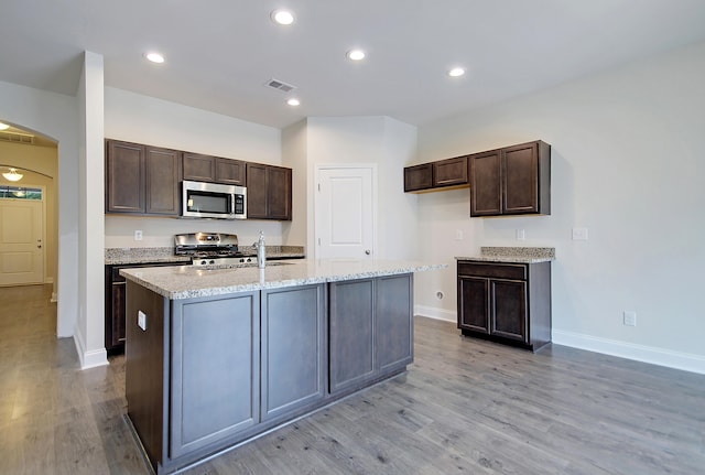 kitchen with stove, light wood-type flooring, an island with sink, dark brown cabinets, and light stone counters