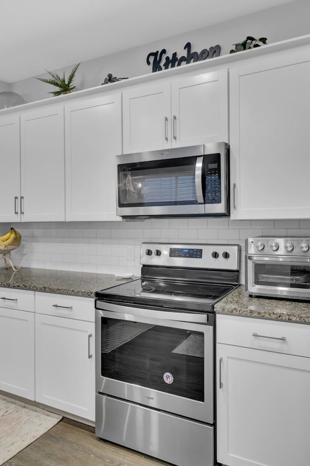 kitchen featuring hardwood / wood-style floors, backsplash, white cabinetry, appliances with stainless steel finishes, and dark stone countertops