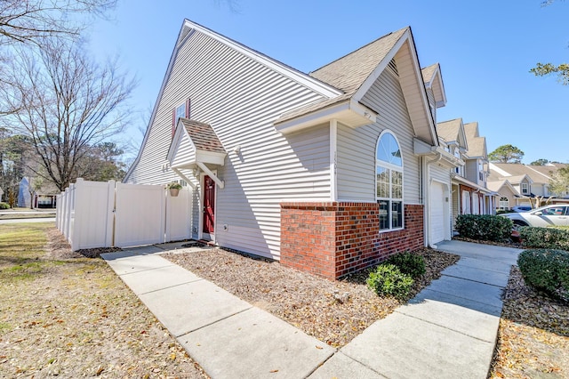 view of home's exterior featuring an attached garage, brick siding, fence, roof with shingles, and a gate