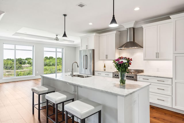 kitchen featuring wall chimney range hood, high end appliances, a tray ceiling, and an island with sink