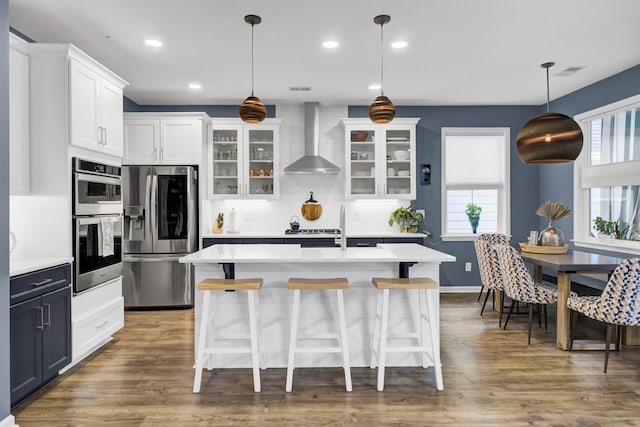 kitchen featuring wall chimney exhaust hood, stainless steel appliances, dark wood-type flooring, white cabinets, and hanging light fixtures