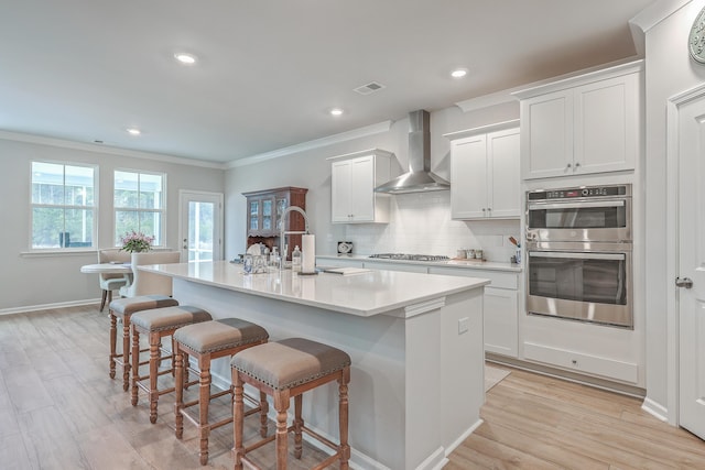 kitchen featuring a center island with sink, wall chimney range hood, stainless steel appliances, decorative backsplash, and white cabinets