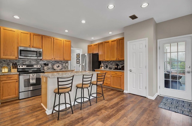 kitchen featuring light stone countertops, stainless steel appliances, a kitchen island with sink, dark wood-type flooring, and a breakfast bar area