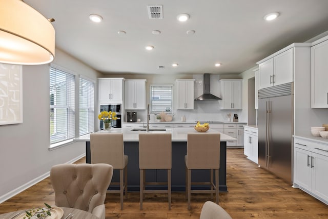 kitchen with built in refrigerator, white cabinets, an island with sink, dark hardwood / wood-style floors, and wall chimney range hood