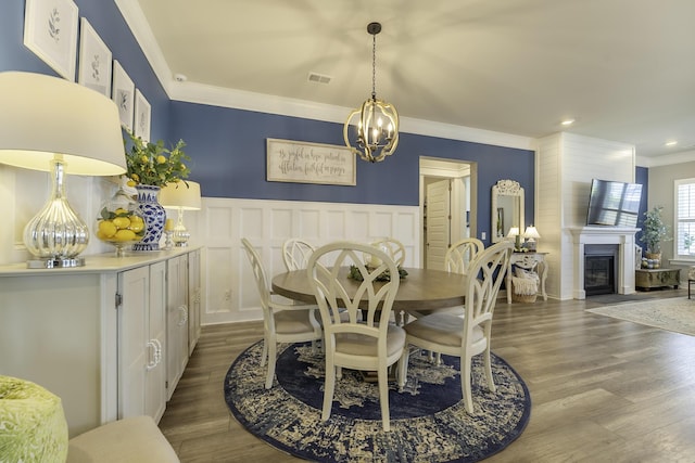dining room featuring ornamental molding, a chandelier, wood finished floors, a glass covered fireplace, and a wainscoted wall