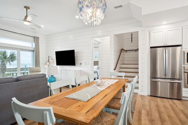 dining space with visible vents, stairs, wood walls, light wood-style floors, and ceiling fan with notable chandelier