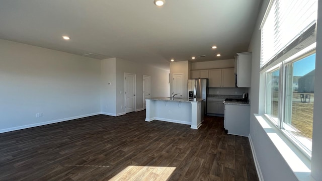 kitchen with dark wood-style floors, stainless steel fridge with ice dispenser, a kitchen island with sink, light stone countertops, and baseboards