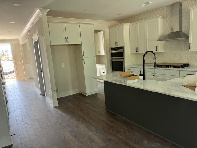 kitchen featuring light stone counters, dark wood-style floors, stainless steel double oven, a sink, and wall chimney exhaust hood