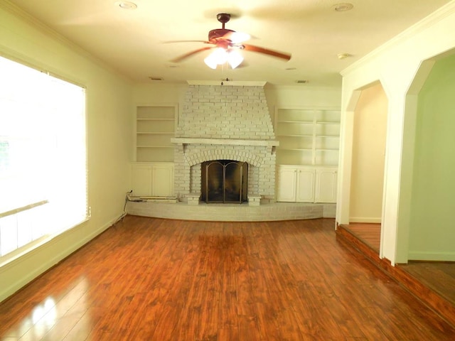 unfurnished living room featuring a brick fireplace, built in shelves, ceiling fan, wood-type flooring, and ornamental molding