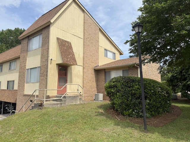 exterior space with cooling unit, a front yard, a shingled roof, and brick siding