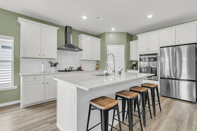 kitchen featuring stainless steel appliances, wall chimney range hood, a sink, light wood finished floors, and a kitchen breakfast bar
