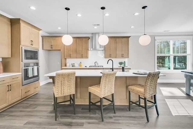 kitchen with pendant lighting, wall chimney range hood, black microwave, an island with sink, and stainless steel oven