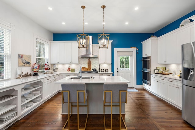 kitchen with wall chimney exhaust hood, stainless steel appliances, a kitchen island with sink, white cabinets, and a breakfast bar area