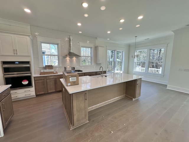 kitchen with pendant lighting, wall chimney range hood, crown molding, wood-type flooring, and a center island with sink