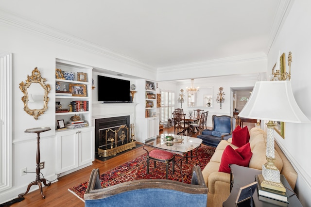 living room with crown molding, built in shelves, wood finished floors, a fireplace with raised hearth, and a chandelier