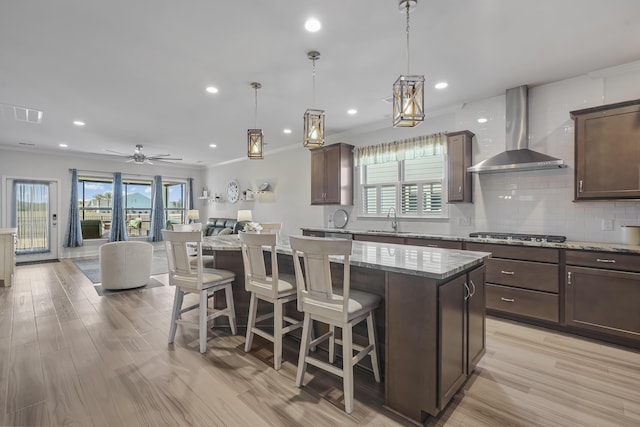 kitchen with stainless steel gas stovetop, open floor plan, a kitchen island, a sink, and wall chimney exhaust hood