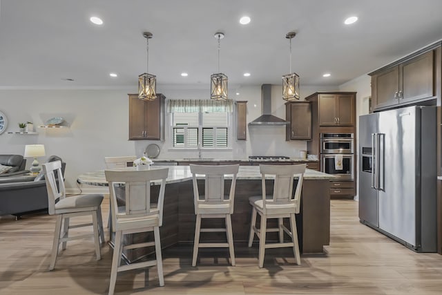 kitchen featuring dark brown cabinetry, appliances with stainless steel finishes, wall chimney range hood, light stone countertops, and a kitchen bar