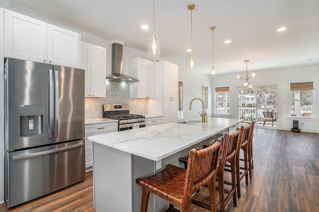 kitchen with hanging light fixtures, appliances with stainless steel finishes, wall chimney range hood, a kitchen island with sink, and white cabinets