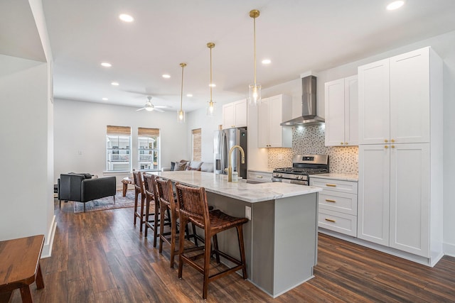 kitchen with white cabinetry, hanging light fixtures, appliances with stainless steel finishes, an island with sink, and wall chimney range hood