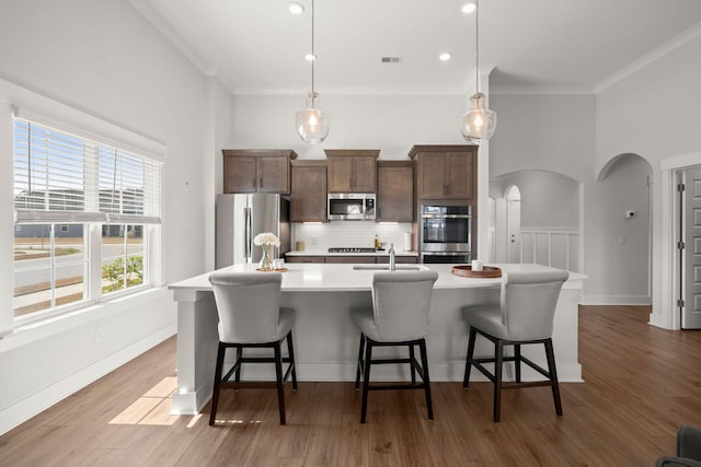 kitchen featuring arched walkways, an island with sink, appliances with stainless steel finishes, wood finished floors, and light countertops