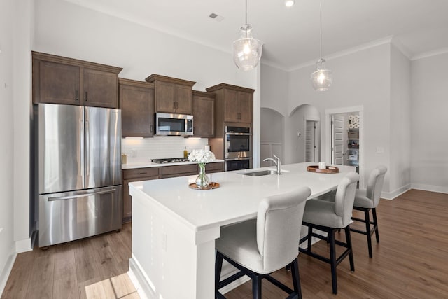 kitchen featuring arched walkways, decorative backsplash, appliances with stainless steel finishes, a kitchen island with sink, and a sink