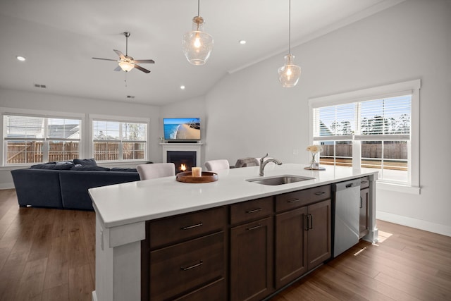 kitchen featuring stainless steel dishwasher, a sink, an island with sink, wood finished floors, and a lit fireplace