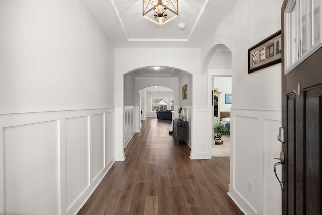 hall featuring dark wood-style floors, a tray ceiling, arched walkways, a decorative wall, and an inviting chandelier