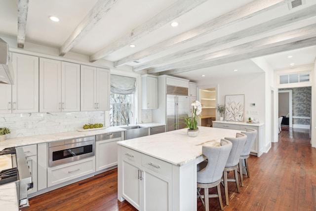 kitchen with a center island, beam ceiling, dark wood finished floors, white cabinetry, and built in appliances