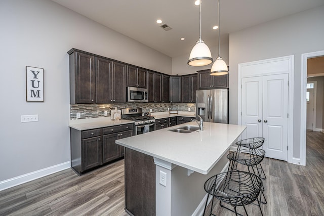 kitchen with dark brown cabinets, hanging light fixtures, an island with sink, stainless steel appliances, and decorative backsplash