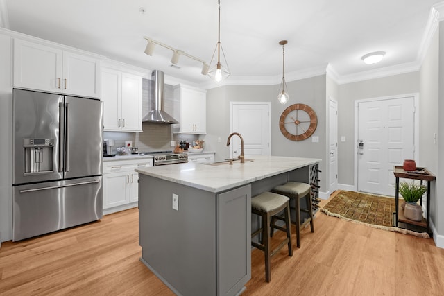 kitchen with white cabinets, an island with sink, sink, wall chimney range hood, and appliances with stainless steel finishes