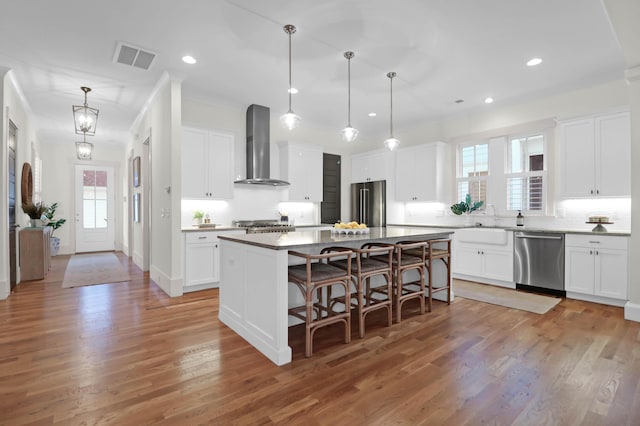 kitchen with appliances with stainless steel finishes, wall chimney range hood, white cabinets, a kitchen island, and hanging light fixtures